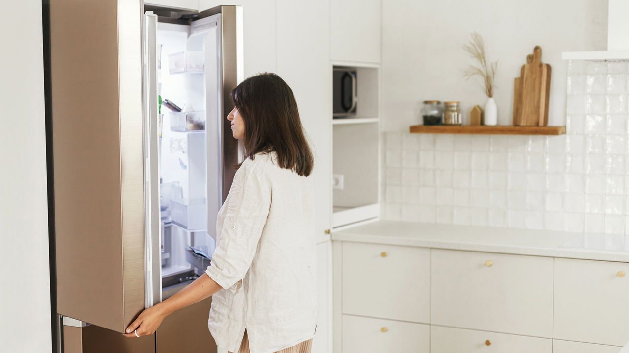 Stylish woman opening fridge and looking inside in new minimal white kitchen Housewife cleaning up
